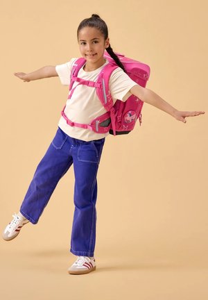 Young girl balancing on one leg, wearing blue pants, white sneakers, beige shirt, and pink backpack with stars and unicorn design.
