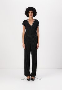 Svart strikket jumpsuit med gullfargede vertikale striper, V-hals, korte ermer, elastisk liv, og avsmalnende bukseben som er samlet ved kanten.