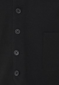 Schwarze Hemd mit Knopfleiste und fünf passenden Knöpfen, strukturierter Stoff und einer einzelnen Brusttasche auf der linken Seite.