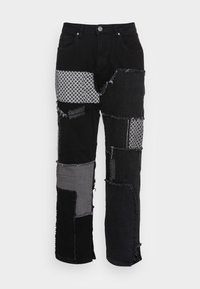 Vaqueros de parches negros con diversos parches de tela en negro y gris, bordes deshilachados y detalles de costura blanca en las piernas delanteras.