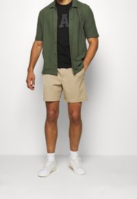 Camisa de botones de manga corta verde sobre una camiseta negra con una letra 'A' gris, combinada con pantalones cortos beige y zapatillas blancas.