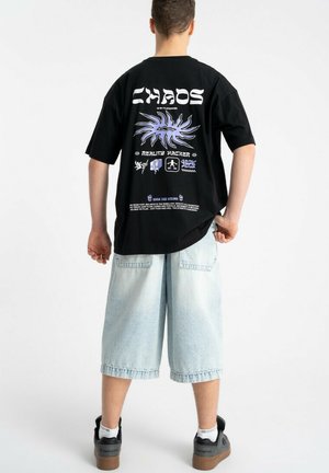 Młoda osoba nosząca czarną koszulkę z nadrukiem "CHAOS" i wzorami, jasnoniebieskie długie szorty jeansowe, białe skarpetki i czarne trampki, stoi tyłem.