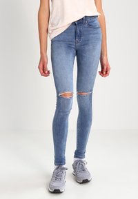Jeans ajustados de color azul claro con detalles de rotura en las rodillas, fabricados en denim. Combinados con zapatillas deportivas grises y una parte superior de color claro.