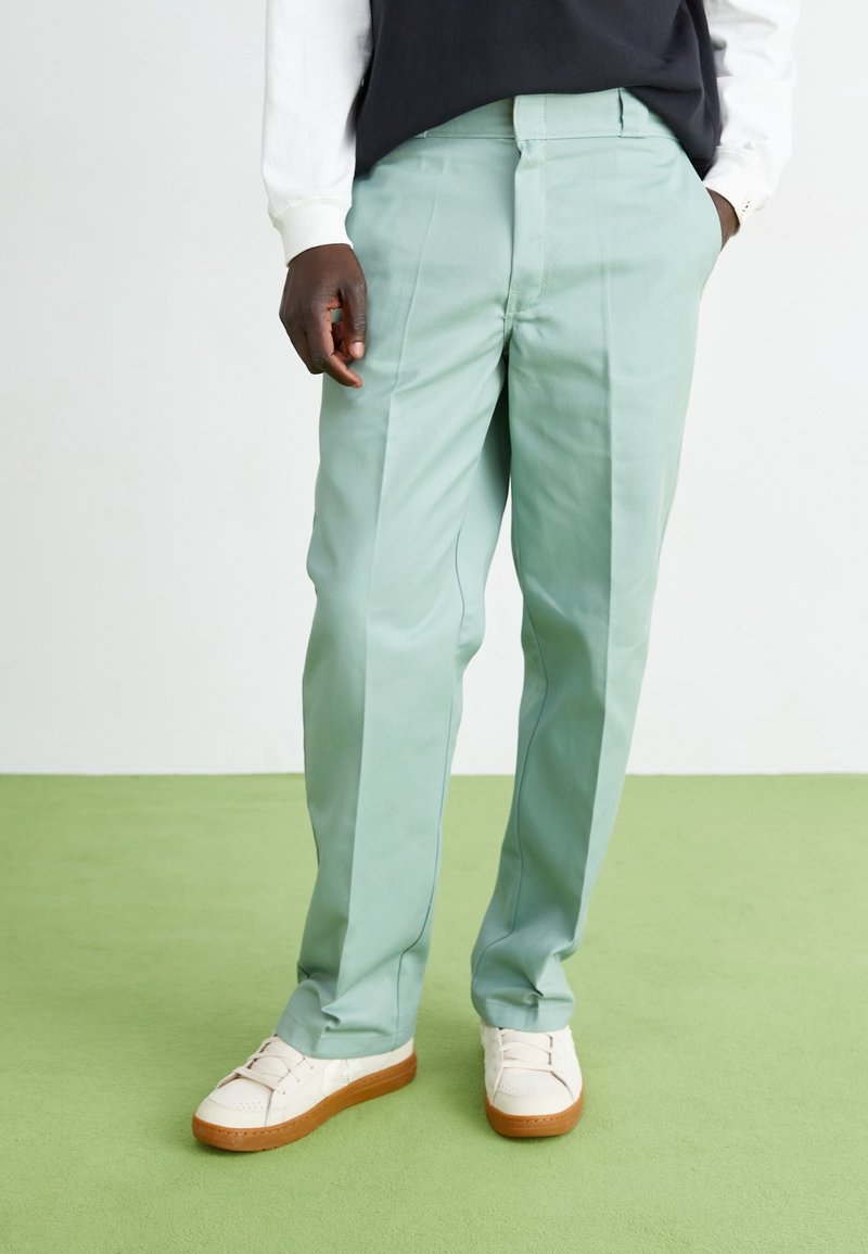 Lichtgroene broek met een getailleerde pasvorm en voorplooi, gemaakt van gladde stof; gecombineerd met witte sneakers.