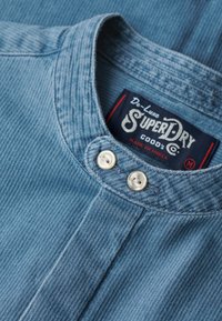 Gros plan d’un col en denim bleu avec deux boutons blancs et une étiquette de marque Superdry noire à l’intérieur, taille moyenne, fabriqué en Inde.