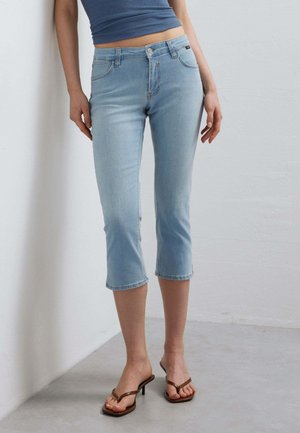 Donna che indossa jeans corti azzurri, una canotta aderente blu senza maniche e sandali marroni con tacco, in piedi contro un muro bianco.