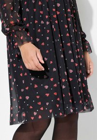 Schwarzes Kleid aus transparenter Stoff, mit einem Muster aus roten Herzen und weißen Pünktchen. Lange Ärmel mit elastischen Bündchen, plissierter Saum.