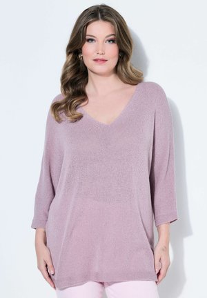 Kvinde med bølget brunt hår iført en lyse lilla V-hals striktrøje og lyserøde bukser, stående mod en ensfarvet hvid baggrund.