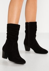 Bottes mi-mollet en suède noir avec des talons bloc et une fermeture éclair sur le côté, portée sur des jambes nues, debout sur un sol blanc.
