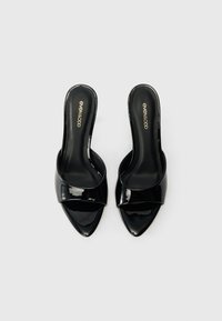 Fekete lakkozott bőr slingback mules, hegyes orral és széles pánttal az elülső részen. Simább textúrával és minimalista dizájnnal rendelkezik.