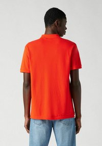 Camisa polo de algodón roja con mangas cortas y cuello clásico, combinada con jeans de mezclilla azul claro. Vista posterior simple sin patrones.