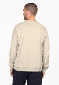 Felpa beige chiaro con collo a girocotolo a coste, maniche lunghe raglan e orlo dritto. Presenta un branding minimale sul lato.
