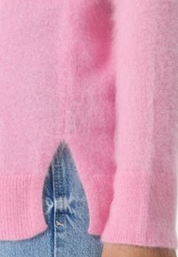 Růžový kašmírový svetr s jemnou, chlupatou strukturou. Má žebrovaný lem a manžety, s bočním rozparkem odhalujícím džíny pod ním.