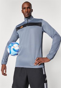 Sudadera deportiva gris de media cremallera con un acento negro en el pecho y detalles de logo naranja, combinada con pantalones cortos negros. Balón de fútbol sostenido en el brazo izquierdo.