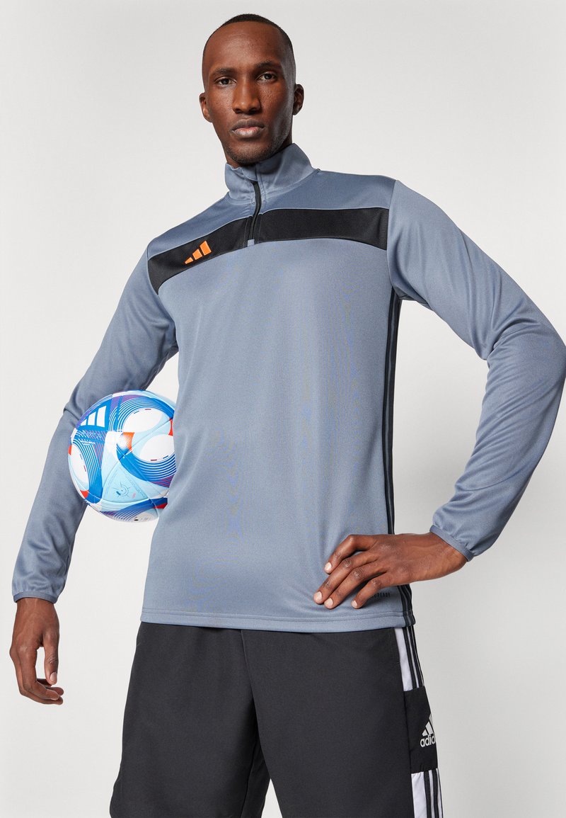 Maglia grigia sportiva a mezza zip con un accento nero sulla parte del petto e dettagli del logo arancioni, abbinata a pantaloni corti neri. Palla da calcio tenuta con il braccio sinistro.