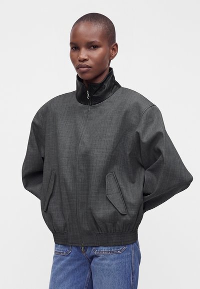 Personne avec des cheveux courts portant une veste zippée ample gris foncé avec un col noir et un jean bleu sur un fond clair uni.