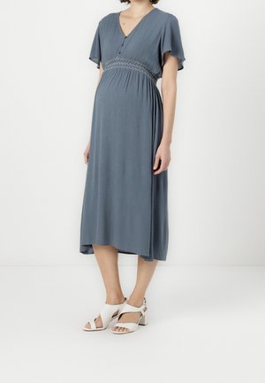 Femme enceinte portant une robe midi bleue avec des manches volantées et des sandales blanches à talons, debout devant un fond blanc.