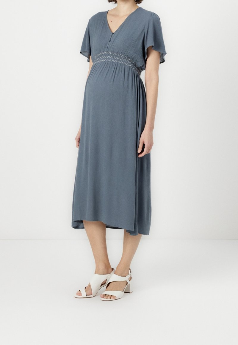 Femme enceinte portant une robe midi bleue avec des manches volantées et des sandales blanches à talons, debout devant un fond blanc.