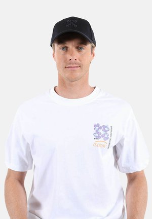 Jeune homme portant un T-shirt blanc avec un logo floral violet et un texte orange, ainsi qu'une casquette de baseball noire portée vers l'avant.
