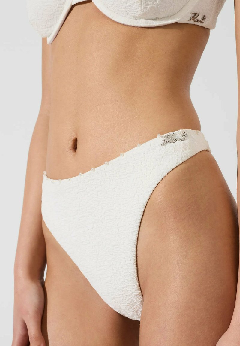 Bas de bikini blanc texturé taille haute et haut assorti portés par une personne, avec de petits détails en perles et un logo métallique "Karl" sur les deux pièces.