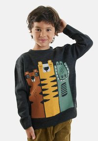 Bambino con capelli ricci corti indossa un maglione scuro con grafiche di un orso dei cartoni animati, una tigre e un dinosauro, abbinato a pantaloni in corduroy marroni.