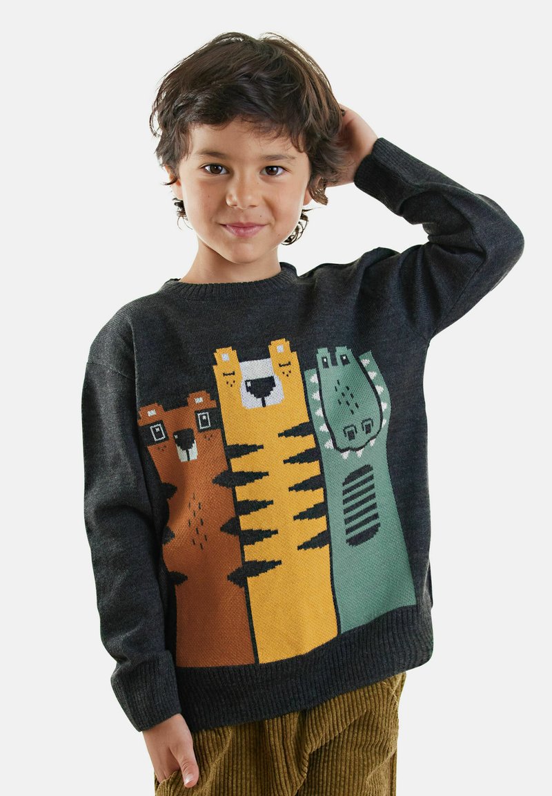 Bambino con capelli ricci corti indossa un maglione scuro con grafiche di un orso dei cartoni animati, una tigre e un dinosauro, abbinato a pantaloni in corduroy marroni.
