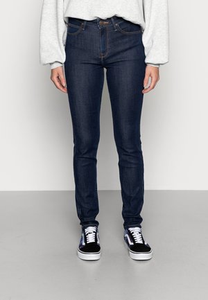 Jeans skinny azul oscuro con costuras naranjas, bolsillos frontales y cierre de botón, combinados con zapatillas negras y blancas.