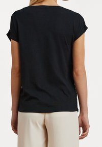 Černé tričko s krátkým rukávem, volného střihu, jemné tkaniny a s ohrnutými rukávy; kombinováno se světlými kalhotami.