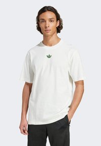 White cotton t-shirt with short sleeves, featuring a green graphic logo on the chest. Paired with black pants, plain design.