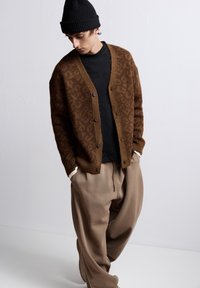 Brun, mönstrad cardigan med knappar, bärs över en svart skjorta. Kombineras med löst sittande, beige byxor med krämfärgade manschetter och en svart mössa.