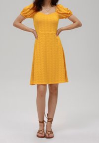Robe jaune avec manches courtes bouffantes, encolure carrée, texture perforée et jupe évasée. Associée à des sandales marron à lanières.