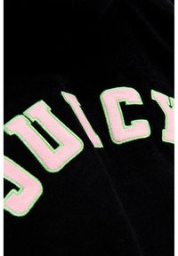 Il tessuto di velluto nero presenta grandi lettere ricamate e soffici con la scritta "JUICY" in rosa e contorno verde, mettendo in evidenza un design con texture leggermente in rilievo.
