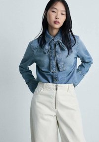 Camicia in denim con dettaglio al collo a cravatta, di colore azzurro chiaro, e chiusura con bottoni, abbinata a pantaloni crema a vita alta con zip metallica.
