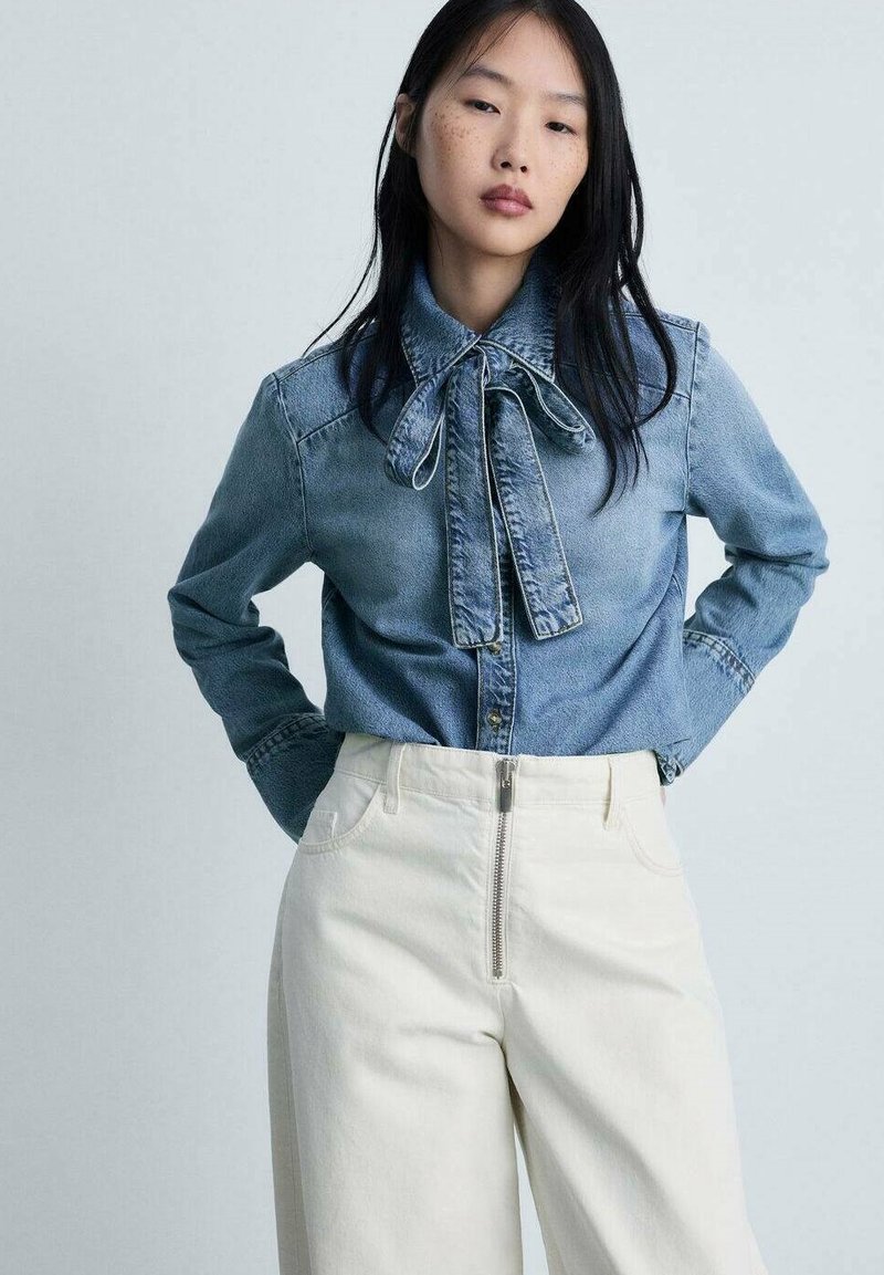 Camicia in denim con dettaglio al collo a cravatta, di colore azzurro chiaro, e chiusura con bottoni, abbinata a pantaloni crema a vita alta con zip metallica.