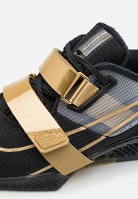Scarpa atletica nera con dettagli in oro, caratterizzata da una tomaia in rete, lacci piatti, una larga fascia piatta e dettagli testurizzati sulla fascia.