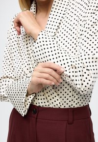 Bluza s točkicama u krem boji s crnim točkama, s širokim manšetama i slobodnim krojem. Uparen s visokim ženskim hlačama u boji burgundca.