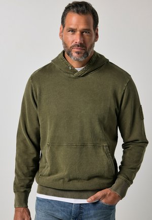 Mann mit kurzen dunklen Haaren und Bart, trägt ein olivgrünes Kapuzensweatshirt mit Fronttasche und blaue Jeans, steht vor einem schlichten Hintergrund.