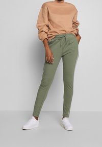 Hellbrauner Sweatshirt mit übergroßen Ärmeln, kombiniert mit enganliegenden grünen Jogginghosen mit weißen Seitenstreifen und weißen Sneakers.