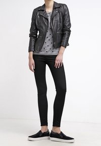 Personne portant une veste en cuir noire, une chemise grise à pois, un pantalon skinny noir et des chaussures noires sans lacets, sur fond blanc uni.