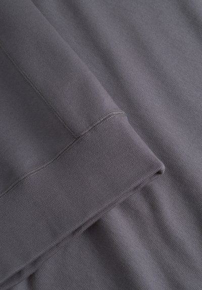 Grauer Stoff mit glatter Textur, ausgestattet mit einem gerippten Bündchen und Nähdetails. Das Material wirkt weich und eignet sich für legere Bekleidung.
