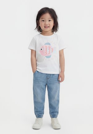 Niño pequeño de pie, viste una camiseta blanca con un dibujo de peces rosa y azul, vaqueros azul claro y zapatillas blancas, sonriendo a la cámara.