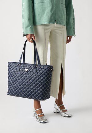 Saco tote azul marinho acolchoado com costura diagonal, alças e acessórios metálicos. Combinado com uma saia branca dividida e sapatos de salto bloco metálico prata.