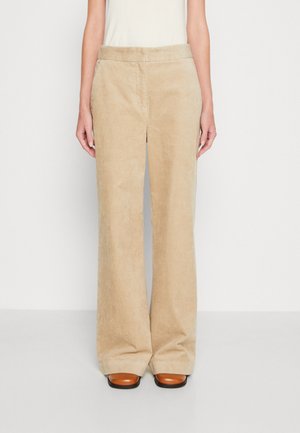 Beige Cordhose mit weitem Bein, kombiniert mit braunen Schuhen mit quadratischer Spitze und einem weißen ärmellosen Oberteil vor neutralem Hintergrund.