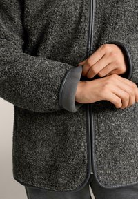 Siva flis jakna z teksturirano površino, kontrastnimi gladkimi manšetami in zadrgo. Rob je zaobljen za sproščen kroj.