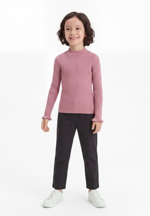Enfant aux cheveux courts et bouclés portant un haut à manches longues côtelé rose, un pantalon noir et des baskets blanches, debout et souriant.