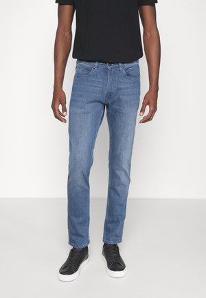 Mann trägt Slim-Fit blaue Jeans, schwarzes T-Shirt und schwarze Sneakers mit weißen Sohlen, steht vor einem einfarbigen weißen Hintergrund.