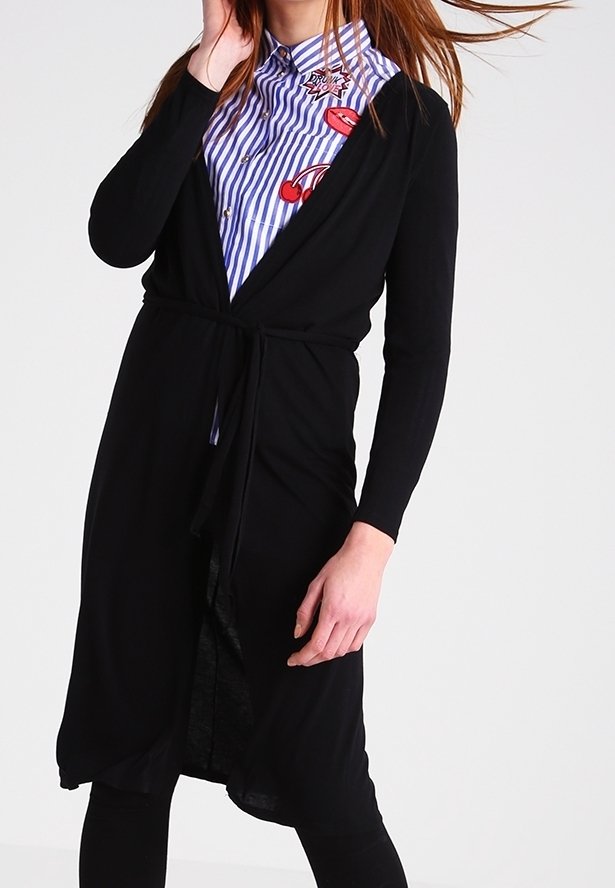 Femme portant un long cardigan noir noué à la taille sur une chemise rayée bleue avec des patchs graphiques rouges sur fond blanc.