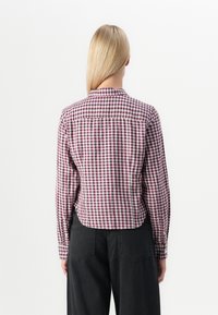 Checked shirt with a maroon and white pattern, long sleeves, buttoned collar, and a slightly slim fit. Dark, high-waisted pants complete the look.