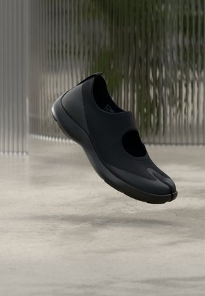 Czarny but wsuwany z rozdzielonym palcem, unoszący się nad gładką betonową podłogą w minimalistycznym wnętrzu.