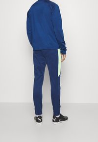 Navy trainingspak met een getailleerd ontwerp, voorzien van een lichtgroene paneel aan de zijkant van de broek. Materiaal lijkt glad, met elastische boorden en zwarte schoenen.